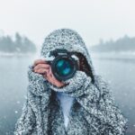 Fotograf i snöfall håller en Nikon-kamera vid en isig sjö